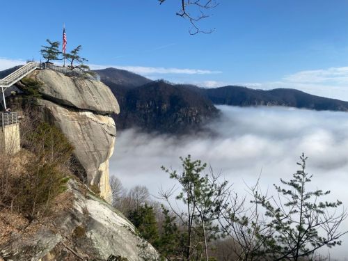 A rocky cliff with a flag overlooks a valley shrouded in fog, surrounded by mountains under a clear blue sky.