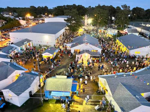 Aerial view of a bustling fairground at dusk, with crowds and colorful tents illuminated by lights.