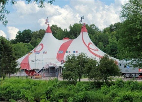 A large red and white circus tent surrounded by trees and greenery under a partly cloudy sky.