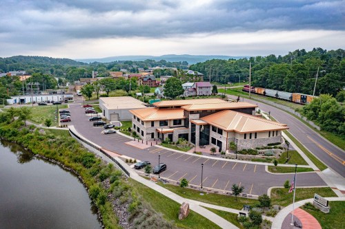 Aerial view of a modern building with a copper roof, surrounded by greenery and a river, near a small town.