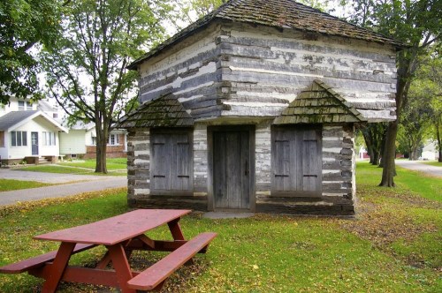 A small, rustic log cabin with a sloped roof, surrounded by trees and a red picnic table in front.