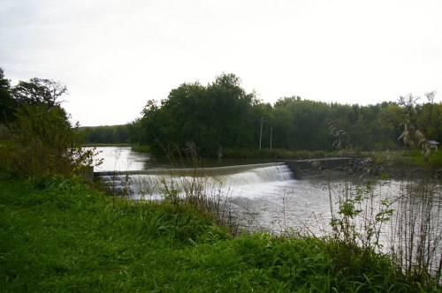 A serene river scene featuring a small waterfall, surrounded by lush greenery and trees under a cloudy sky.