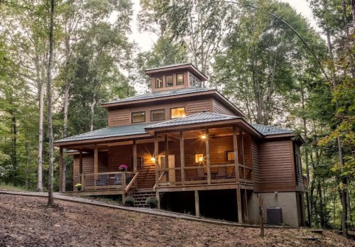 A large wooden house with a porch, surrounded by trees, featuring a unique tower and warm lighting at dusk.