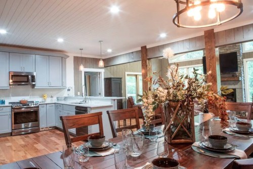 A modern kitchen and dining area with wooden accents, a large table, and a floral centerpiece. Bright and inviting atmosphere.