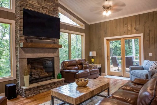 Cozy living room with a stone fireplace, large windows, and comfortable seating, overlooking a wooded area.