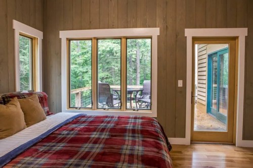 Cozy bedroom with a plaid blanket, large windows overlooking a forest, and a door leading to a deck.