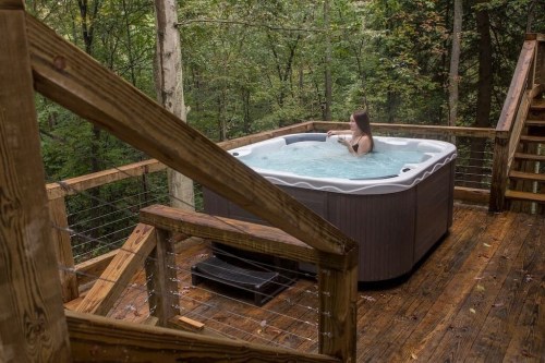 A person relaxes in a hot tub on a wooden deck surrounded by lush green trees.