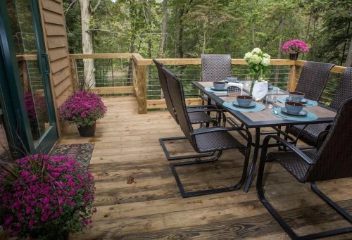 A cozy outdoor dining area with a table set for four, surrounded by potted flowers and a lush forest backdrop.