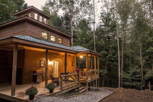 A cozy wooden cabin with a porch, surrounded by trees, in a serene forest setting at dusk.