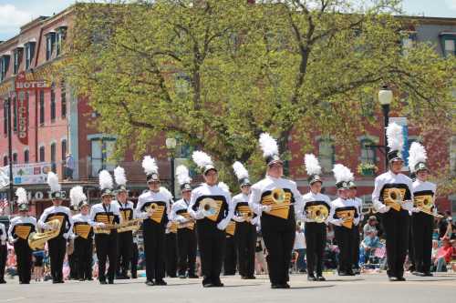 A marching band in black and white uniforms parades down a street, with spectators and buildings in the background.