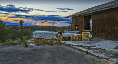 A rustic outdoor space with hay bales, a wooden structure, and a colorful sunset over distant mountains.