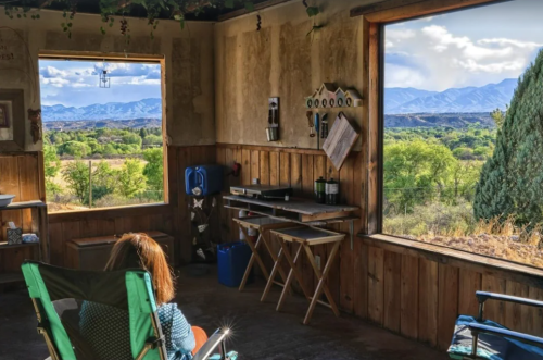 A person sits in a rustic room, gazing out at a scenic view of mountains and greenery through large windows.