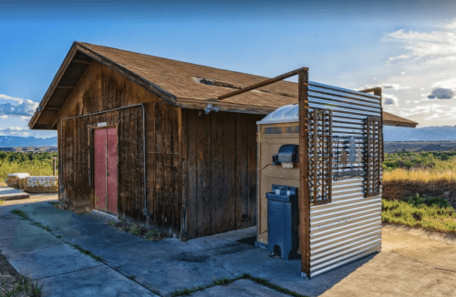 A rustic wooden building with a metal addition, set against a backdrop of mountains and a blue sky.