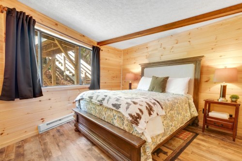 Cozy bedroom with wooden walls, a queen bed, two lamps, and a window overlooking a natural setting.
