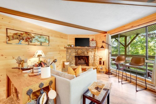 Cozy living room with wooden walls, a stone fireplace, and a view of nature through large windows.