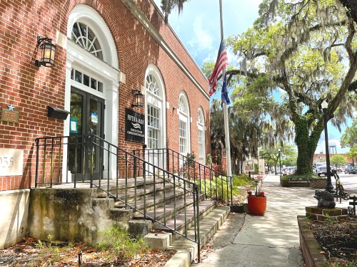 Historic brick building with stairs, American flag, and trees draped in Spanish moss in a sunny outdoor setting.