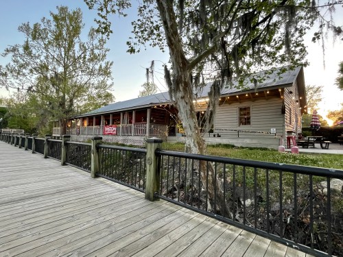 A wooden boardwalk leads to a rustic building with a porch, surrounded by trees and greenery at sunset.