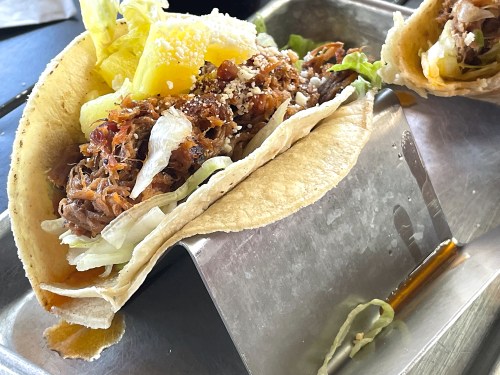 A close-up of a taco filled with shredded meat, lettuce, and pineapple, served on a metal tray.