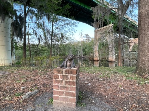A bronze statue of a cat sits on a brick pedestal near a riverbank, with a bridge and trees in the background.
