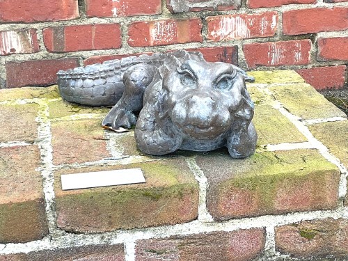 A bronze alligator sculpture resting on a stone surface, with a brick wall in the background.
