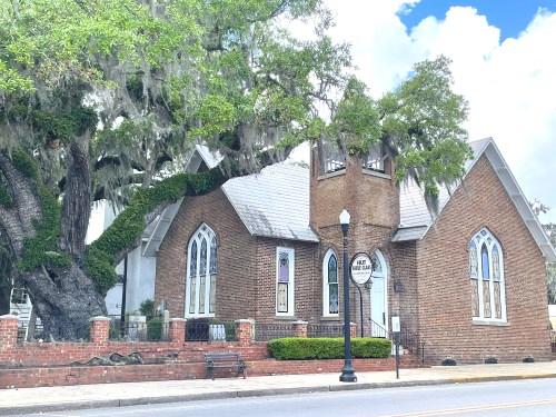 A brick church with large windows, surrounded by greenery and a massive tree, under a blue sky with clouds.
