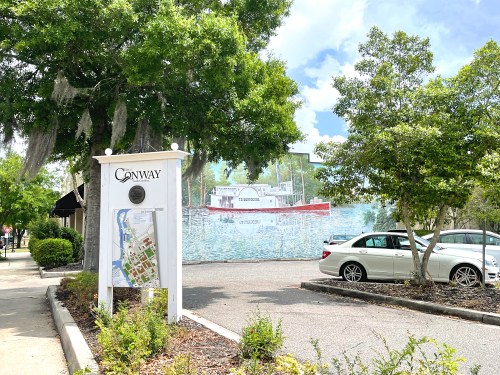 Sign for Conway with a map, surrounded by greenery and a mural of a boat in the background. Cars parked nearby.
