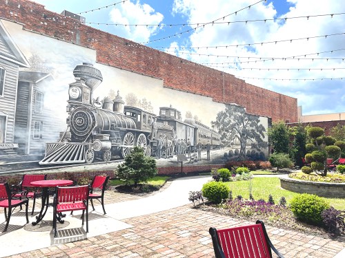 A mural of a vintage steam train on a brick wall, surrounded by a garden with tables and chairs.