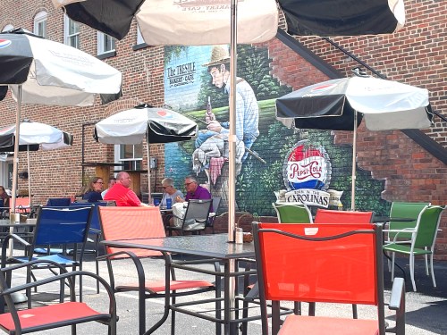 Outdoor dining area with colorful chairs and umbrellas, featuring a mural of a man fishing on a brick wall.