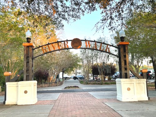 Archway sign reading "River Walk" surrounded by trees and a paved pathway. Soft evening light illuminates the scene.
