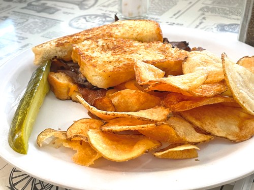 A plate with a grilled cheese sandwich, crispy potato chips, and a pickle on the side.