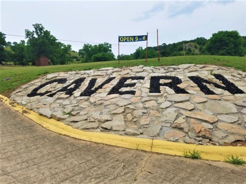 Sign made of stones spelling "CAVERN" on a grassy area, with an "OPEN 9-6" banner in the background.