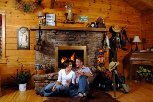 A couple sits by a stone fireplace in a cozy wooden cabin, surrounded by fishing gear and rustic decor.