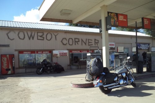 A motorcycle parked outside a rustic gas station with "Cowboy Corner" painted on the wall.