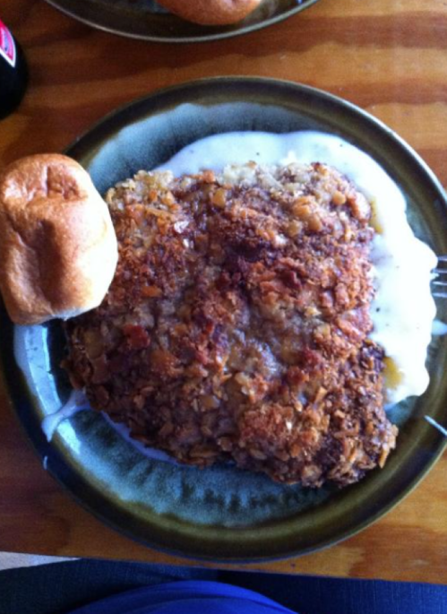 A plate of fried chicken smothered in gravy, served with mashed potatoes and a roll.