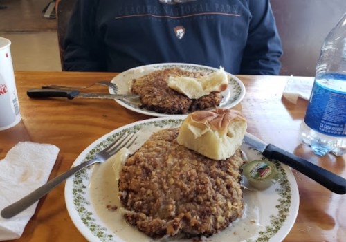 Two plates of food with large, round patties and a bun, set on a wooden table with utensils and a water bottle.
