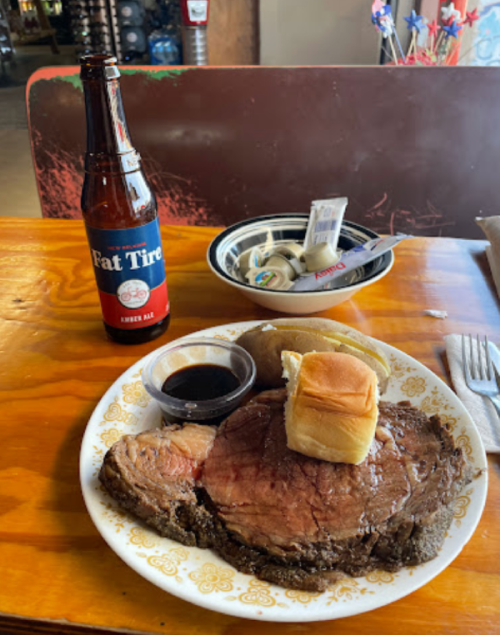 A plate with prime rib, a roll, and dipping sauce, alongside a bottle of Fat Tire beer on a wooden table.