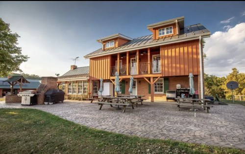 A large, modern orange house with a balcony, surrounded by picnic tables and a grill in a grassy area.