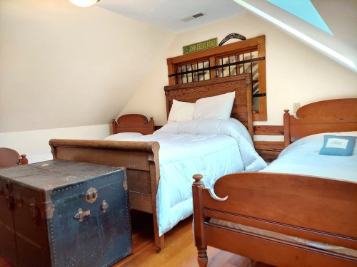 Cozy attic bedroom with two wooden beds, a vintage trunk, and sloped ceiling, featuring warm wood tones and natural light.