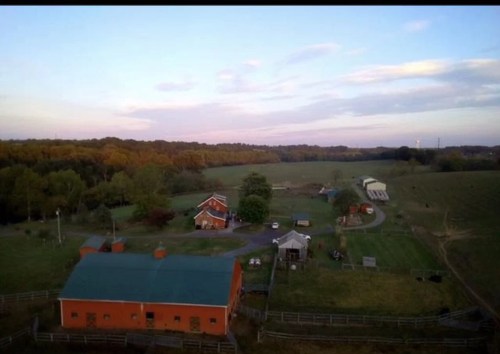 Aerial view of a farm with red buildings, green fields, and trees under a colorful sky at dusk.