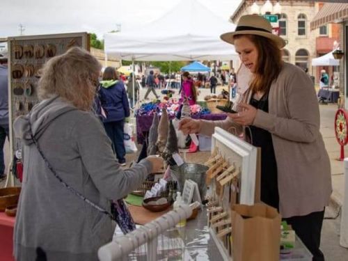 A vendor hands a customer a handmade item at a bustling outdoor market with tents and people in the background.