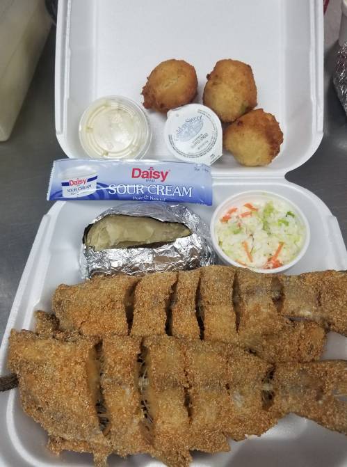 A takeout container with fried fish, a baked potato, coleslaw, sour cream, and three hushpuppies.