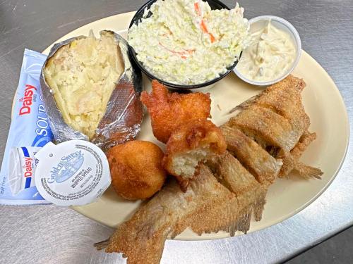 A plate of fried fish, coleslaw, a baked potato, hushpuppies, and dipping sauces.
