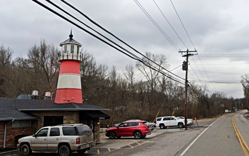 A red and white lighthouse structure beside a road, with parked cars and bare trees in a cloudy sky.
