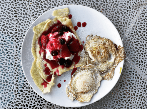 A plate with a crepe topped with berries and cream, alongside two fried eggs, on a patterned table.