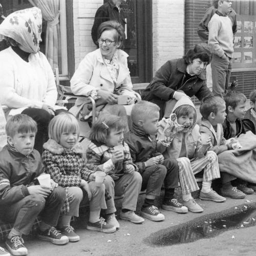 A group of children sitting on a curb, enjoying snacks, with adults in the background, in a vintage black-and-white photo.