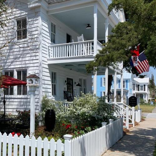 A charming white house with a porch, surrounded by greenery and flowers, featuring American flags and a picket fence.