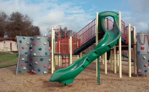 A playground featuring a green slide, climbing wall, and multi-level play structure on a gravel surface.