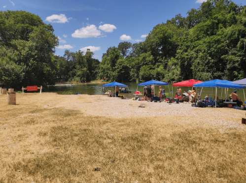 A riverside scene with colorful tents, people relaxing, and lush greenery under a blue sky with scattered clouds.