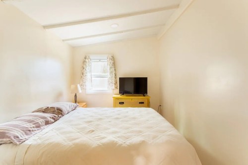 Cozy bedroom with a queen bed, a lamp, a yellow dresser, and a TV by the window. Light-colored walls and natural light.