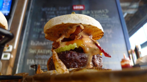 A close-up of a juicy burger with melted cheese, bacon, pickles, and a toasted bun, set against a blurred background.